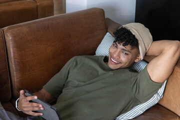 Fototapeta premium African American male reclining on leather sofa at home holding tablet and wearing beanie