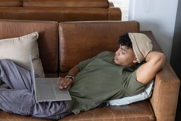 Fototapeta premium African American male reclining on brown leather sectional sofa in living room using silver laptop