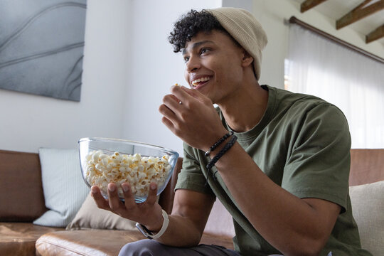 African American man beanie-wearing sitting on sofa at home holding glass bowl eating popcorn