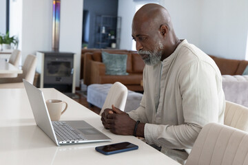 Fototapeta premium Senior man sitting at table, working on silver laptop, phone and ceramic mug, copy space