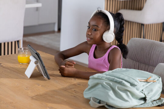 African American teen girl sitting at wood table at home wearing white headphones using tablet