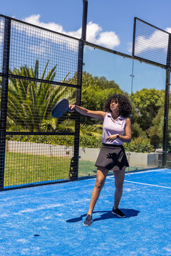 Woman standing on blue court in purple top and black skort holding perforated paddle and smartwatch