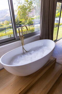 Oval white bathtub is sitting on platform in bathroom near windows with brass faucet and foam