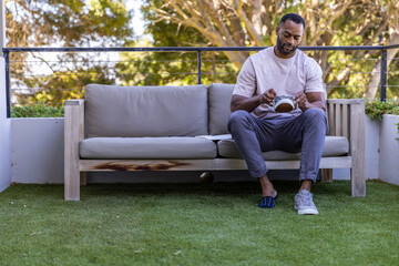 Fototapeta premium Mature African man sitting on right balcony sofa cleaning sneaker with brush and cloth, copy space