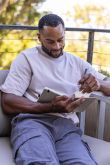 Fototapeta premium Mature man sitting on balcony chair writing in notebook with pen and cargo pants
