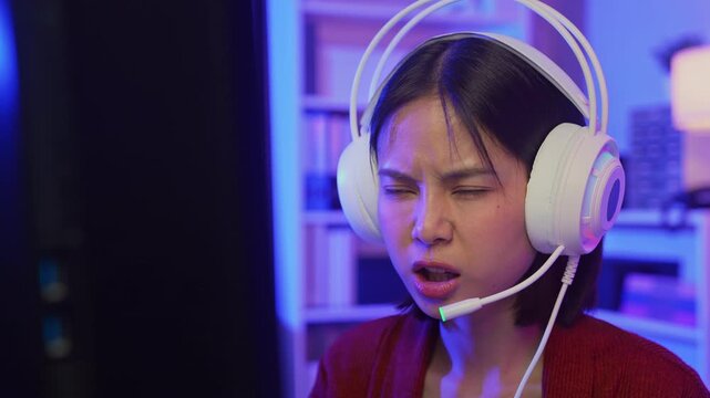 Excited young Asian girl playing online computer game with headset and microphone at home, esports and live streaming concept.