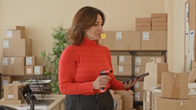 Woman scans cardboard parcels with handheld barcode scanner and taps tablet while organizing packages in a small business building; confidence efficiency.