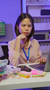 Stressed young Asian office worker feeling overwhelmed with paperwork and workload while working on laptop in office