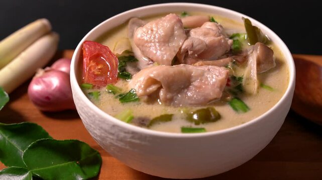 Traditional Thai Tom Kha Gai soup served in a white bowl with chicken, coconut milk, lemongrass, kaffir lime leaves, shallots, and fresh herbs on a wooden table. Authentic Thai cuisine with rich cream