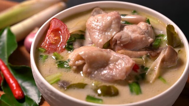 Traditional Thai Tom Kha Gai soup served in a white bowl with chicken, coconut milk, lemongrass, kaffir lime leaves, shallots, and fresh herbs on a wooden table. Authentic Thai cuisine with rich cream