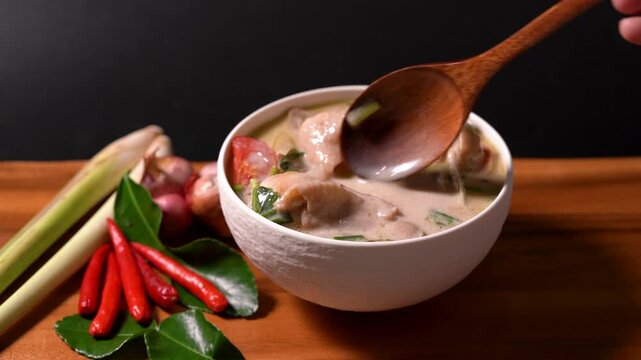 Traditional Thai Tom Kha Gai soup served in a white bowl with chicken, coconut milk, lemongrass, kaffir lime leaves, shallots, and fresh herbs on a wooden table. Authentic Thai cuisine with rich cream