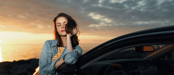 Fototapeta premium Relaxed woman leaning against a car at sunset, denim jacket striped top, wind in hair. A lifestyle moment highlighting travel, freedom, and outdoor evenings.