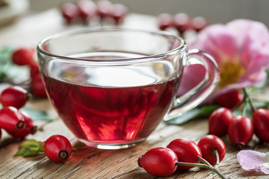 A transparent glass cup filled with red herbal rose hip tea, surrounded by fresh and dried rosehips and pink flowers.