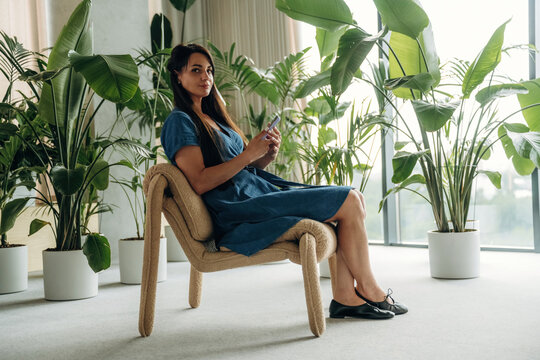 Woman in blue dress sitting on unique chair surrounded by large indoor plants in a modern, bright room with floor-to-ceiling windows and soft natural light