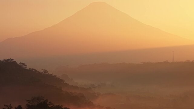 Aerial view of a majestic volcano at sunrise with hazy atmosphere over the lush tropical hills of Selomerto, Central Java, Indonesia.
