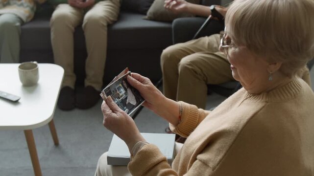 Side view shot of senior woman looking at printed family photographs and smiling in retirement home. Clip for senior care facility advertisements and memory care marketing campaigns