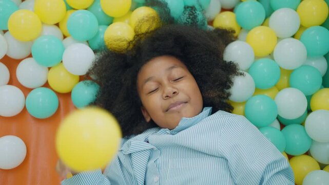 Child's Bliss in a Ball Pit: A young child with an afro relaxes contentedly, surrounded by a vibrant array of colorful balls in a ball pit, embodying pure childhood joy and carefree moments.