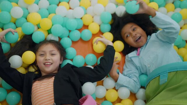 Joyful Kids in Ball Pit: Two happy young girls are having fun playing in a colorful ball pit, surrounded by hundreds of vibrant plastic balls. They are lying on their backs and smiling at the camera.