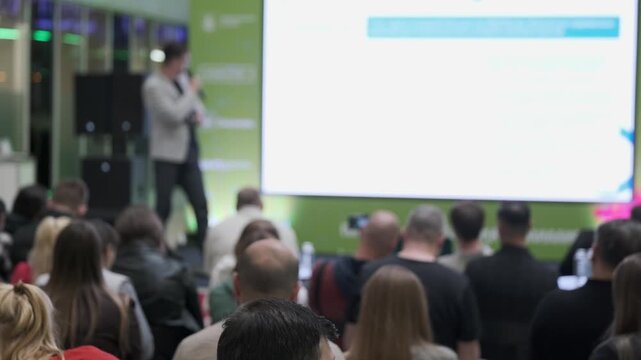 Conference audience watches speaker presenting on large screen. Rows of attendees seen from rear, focus on crowd and event atmosphere.