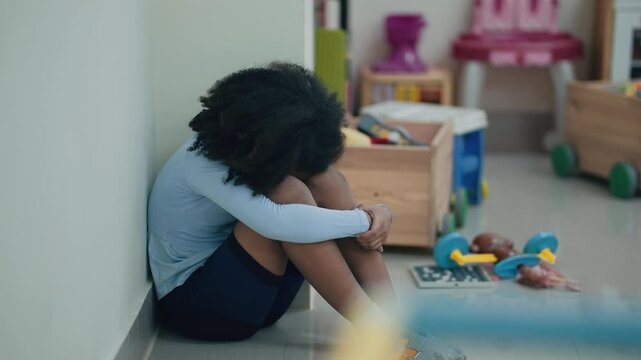 Isolated Child in Playroom: A young child sits alone, head bowed and curled into a ball, in a colorful playroom filled with toys. The scene evokes feelings of sadness, loneliness, and neglect.