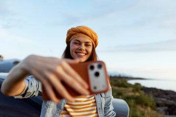 Fototapeta premium Bright lifestyle portrait of a cheerful woman taking a selfie outdoors with a smartphone, wearing a headband and striped shirt, denim jacket beside a coastal road.
