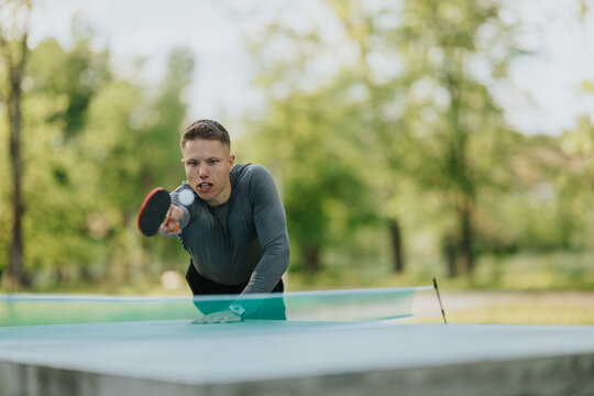 Young male table tennis player reaches forward to return a fast shot during an outdoor match. Action shot shows a focused athlete playing ping pong at a concrete park table.