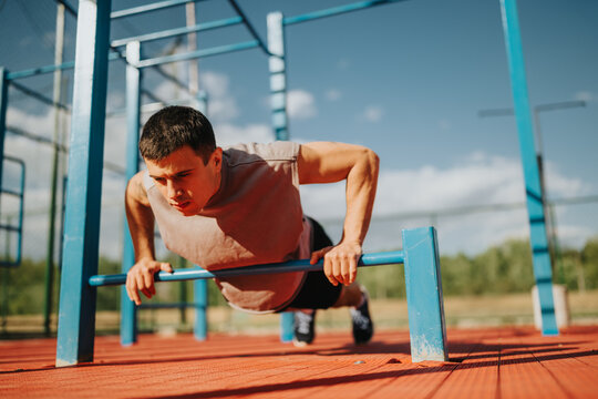 A young man performs push-ups on parallel bars at an outdoor fitness park. He trains with bodyweight calisthenics on a sunny day, demonstrating strength and focus.