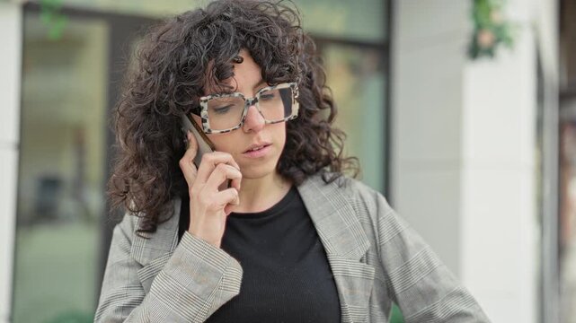 Woman holding smartphone to ear, talking with hand near face and wearing large glasses and plaid blazer outside building storefront in urban street scene; pensive concern.