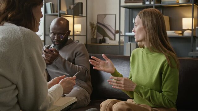 Medium shot of young adult Caucasian woman talking to supportive female psychotherapist during couples therapy session with Black husband. For mental health and counseling services