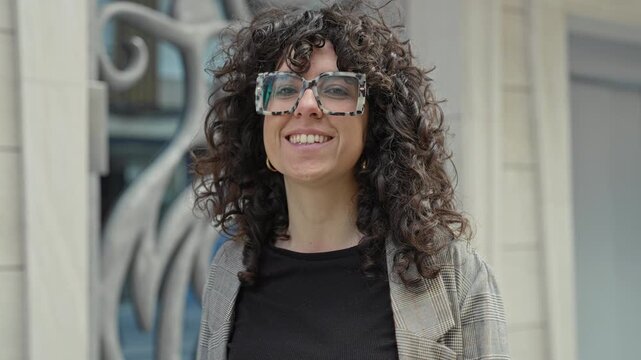 Woman wearing oversized tortoiseshell glasses smiles at camera showing teeth while standing by decorative metal sculpture and stone facade on street; confidence warmth.