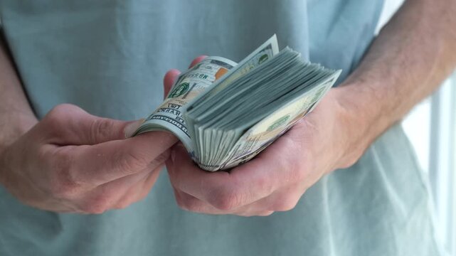 Close-up of hands counting crisp banknotes. Fingers gently roll through a thick stack of currency. Person wears a light-colored, plain shirt.