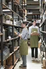 Fototapeta premium A young saleswoman and salesman walk a homeware aisle reviewing stock, the woman noting items on her clipboard while the man scans the bottles, deep among the black shelving