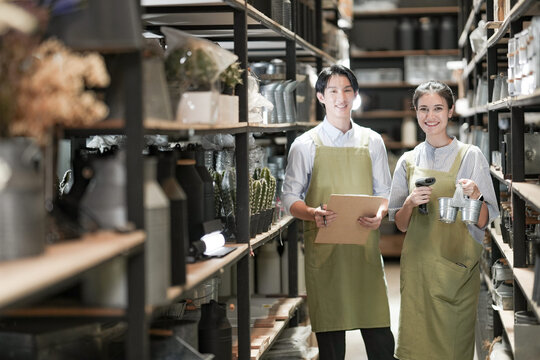 Two young homeware shop owners stand smiling in their stockroom aisle, the saleswoman holding a clipboard and the salesman a small jar, proud of their growing small business