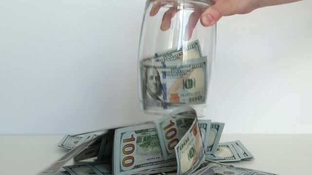 Hand drops hundred-dollar bills into clear glass jar. Cash spills onto white surface below. Money accumulates, symbolizing wealth or savings.