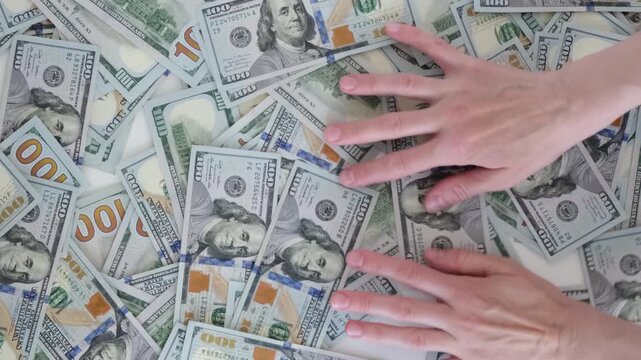 Hands sift through scattered hundred-dollar bills. Fingers trace bills, feeling wealth's abundance. No emotion shown, just focused movement. 