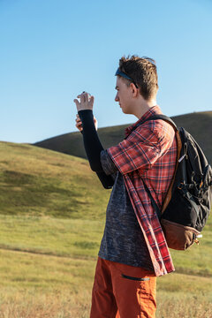 A young teenage boy traveler is standing on a grassy hill in Kyrgyzstan mountains under a clear blue sky, holding a smartphone and taking a picture or filming a video.