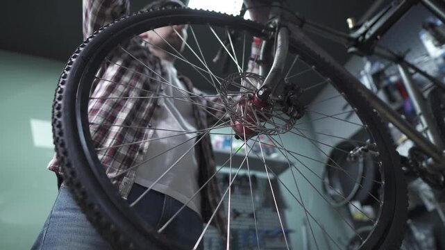 Low angle shot of a male bike mechanic in a plaid shirt repairing and adjusting the front wheel and disc brake system of a mountain bicycle on a special stand inside a professional workshop garage.