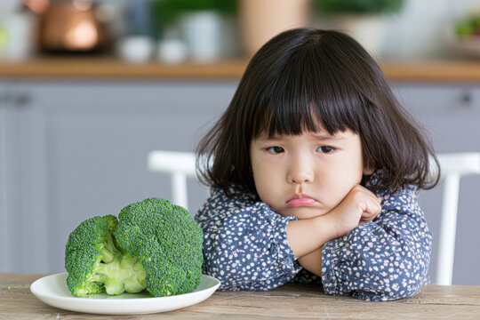 A young girl looks unhappy and disinterested, resting her chin on her hands while staring at a plate of broccoli in front of her.