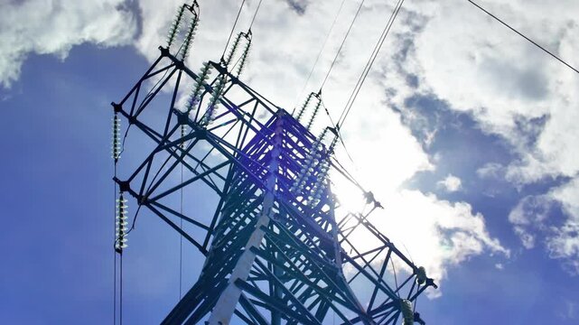 Towering high-voltage electricity pylon with glass insulators silhouetted against a bright sun and blue sky with white clouds, representing the concept of power grid and energy distribution