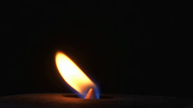 Rightward leaning flame rising from small conical sand wick in dark minimal setting with glowing orange core, soft blur, subtle heat shimmer, and quiet isolated atmosphere