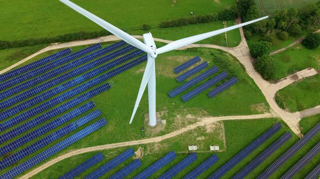 Aerial view of the Chelveston Hybrid Solar and Wind Energy Farm, featuring rows of solar panels and a wind turbine on green fields in Wellingborough, England, United Kingdom.