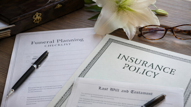 Funeral planning documents with insurance policy, lily and pen on wooden desk. End of life preparation and memorial service paperwork concept.