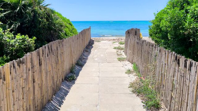 POV walk to the beach in Sardinia along a cemented path surrounded by coastal vegetation leading toward the Mediterranean sea in natural light.