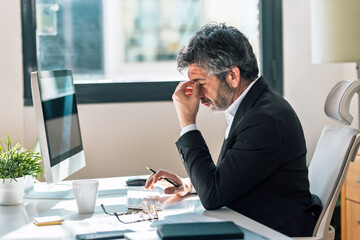 Obraz premium Stressed elegant businessman with a headache working on a computer in the office.