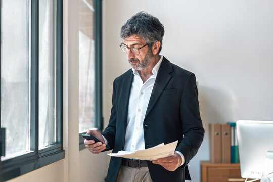 Elegant businessman reading some notes while using smartphone standing  at the office.