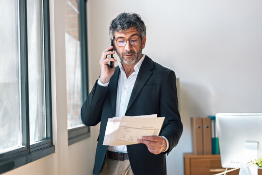 Elegant businessman reading some notes while talking with smartphone standing  at the office.