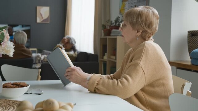 Side view shot of senior woman wearing glasses sitting at table and reading book in retirement home. Clip suitable for promoting senior care services, active aging, and lifelong learning