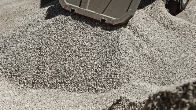 Dump truck unloading crushed stone into rising gravel mound at sunlit industrial yard, rear bed lifted high as heavy material pours out over compacted ground with dusty textures