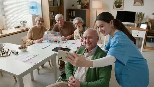 Senior man in wheelchair and young female nurse taking selfie on smartphone in nursing home. Footage marketing geriatric care, assisted living, and nursing services