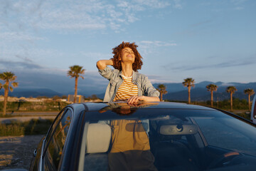 Fototapeta premium Woman leans on car roof under sunny sky, casual denim striped top, open air mood, outdoor lifestyle moment, relaxed and cheerful, bright day, breeze, horizon, nature, palm trees background, candid